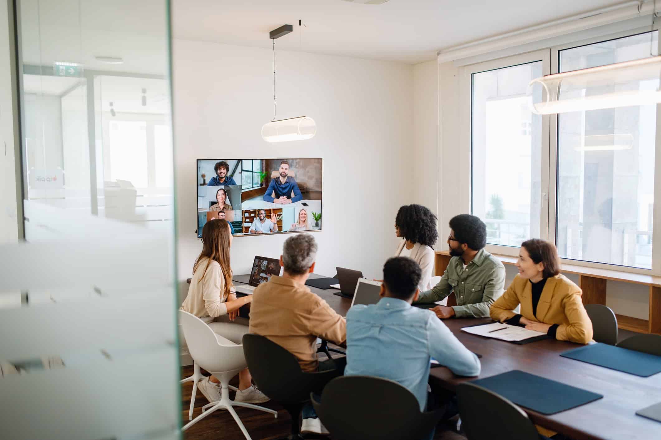 Reunión de equipo en formato híbrido, con empleados conectados por videollamada y otros presentes en la sala, fomentando la comunicación efectiva en el trabajo.