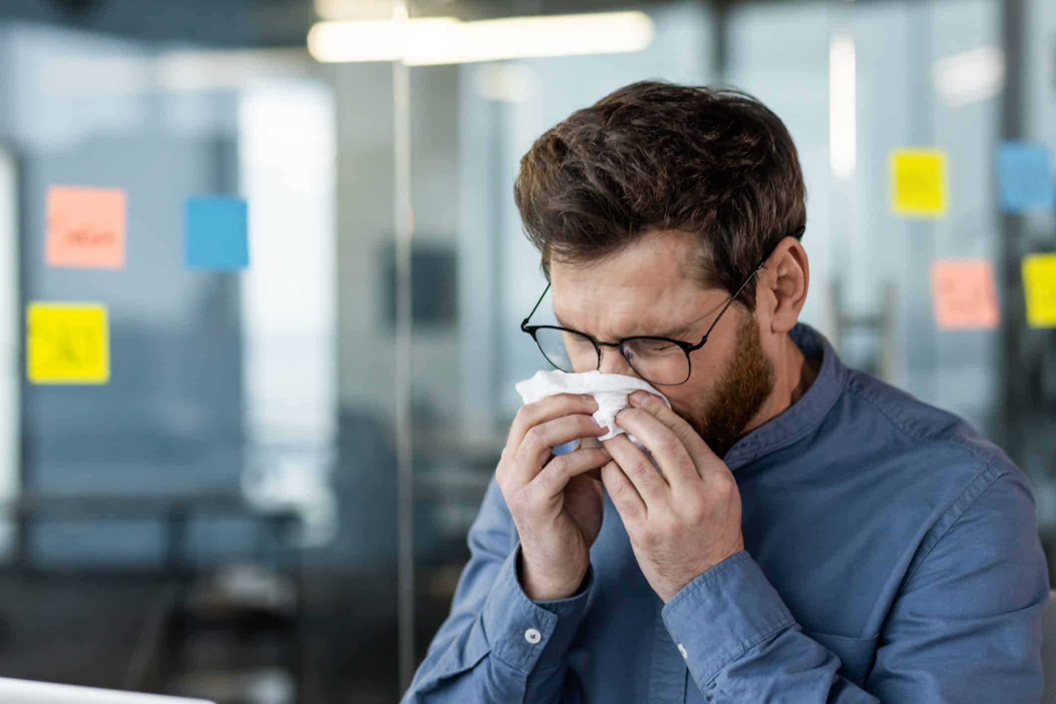 Hombre en una oficina limpiándose la nariz con un pañuelo, síntoma de resfriado o gripe
