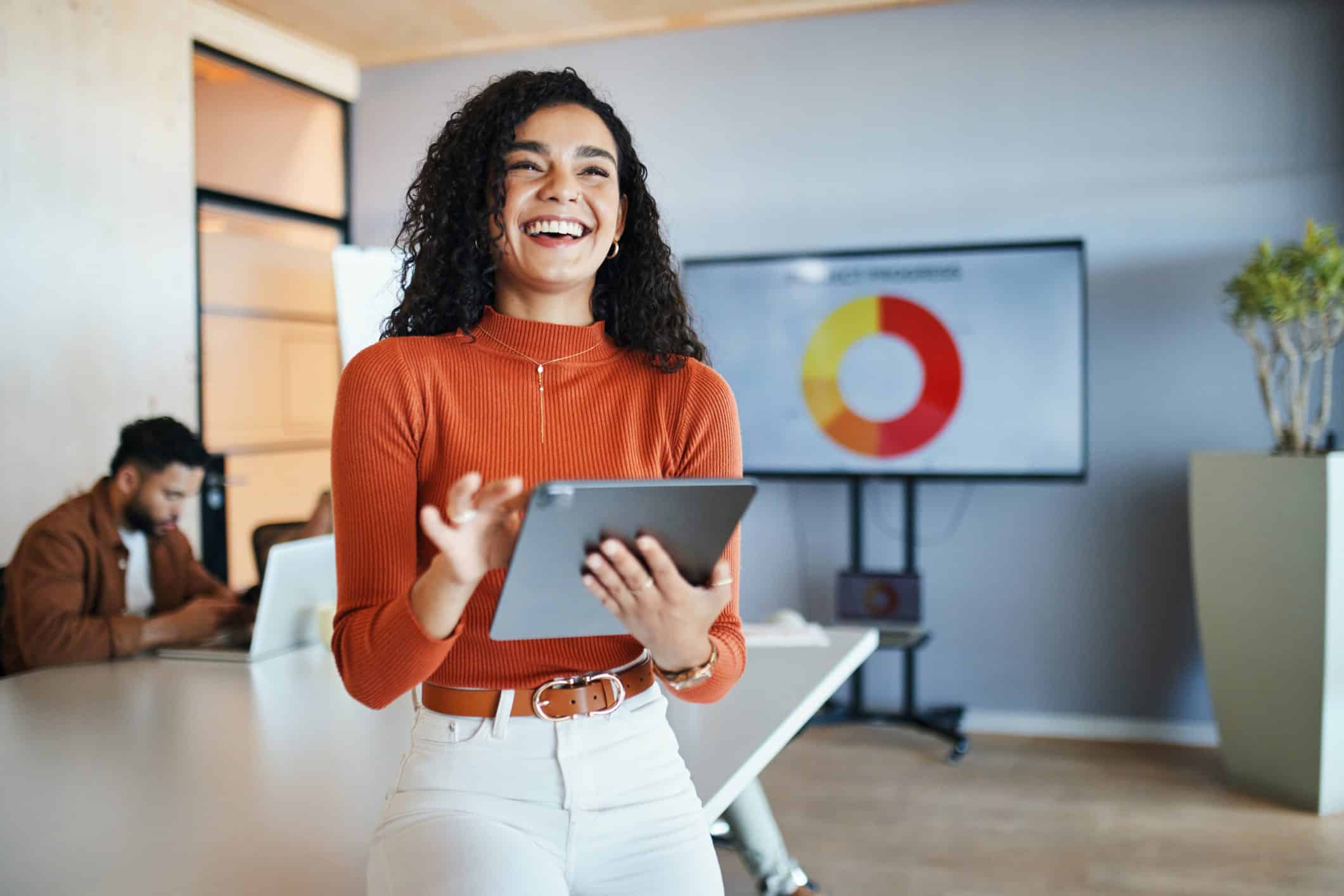 Mujer sonriendo en la oficina con una tablet en la mano, a su espalda, una pantalla con gráficos y datos.