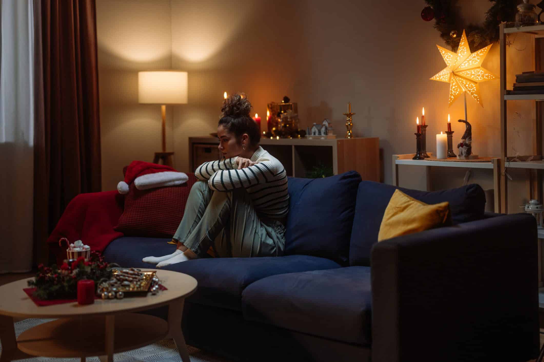 Chica joven sentada sola en el sofá durante la Navidad, mostrando signos de tristeza y soledad en un ambiente navideño iluminado.